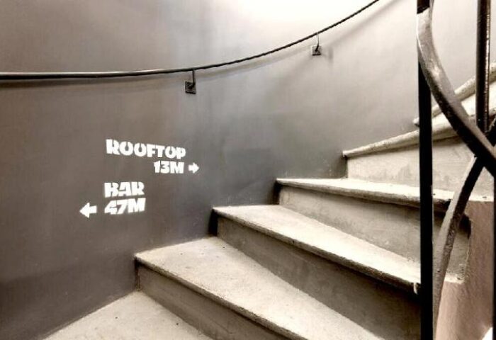 Staircase at Les Piaules, a gay-friendly hotel. Wall signage indicates Rooftop 13M and Bar 47M. Spiral staircase with metal railing.