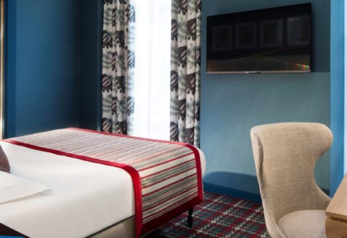 Les Tournelles gay-friendly hotel room. Modern design with blue walls, patterned curtains, and a flat-screen TV. Comfortable chair and stylish bed.