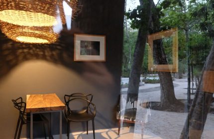 Loba Tulum Male Only gay-friendly hotel interior. Wicker lamp above a table and chairs. View of trees through a window.