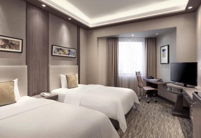 M Hotel gay-friendly hotel room with two beds, desk, and TV. Modern design, neutral colors, and artwork on the walls. Comfortable and inviting.