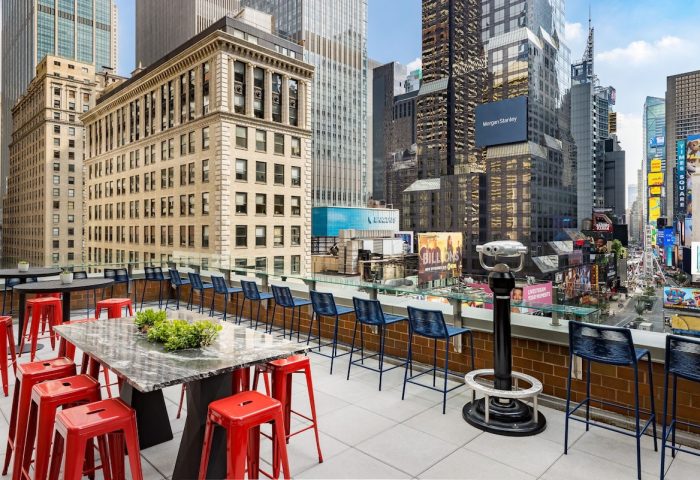 Rooftop view from gay-friendly M Social Times Square hotel. Red stools surround a table. Binoculars overlook Times Square. NYC skyline.