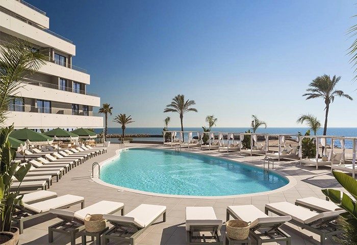 ME Sitges Terramar, a gay-friendly and pet-friendly hotel. Poolside view with lounge chairs, palm trees, and ocean backdrop. Luxury travel.