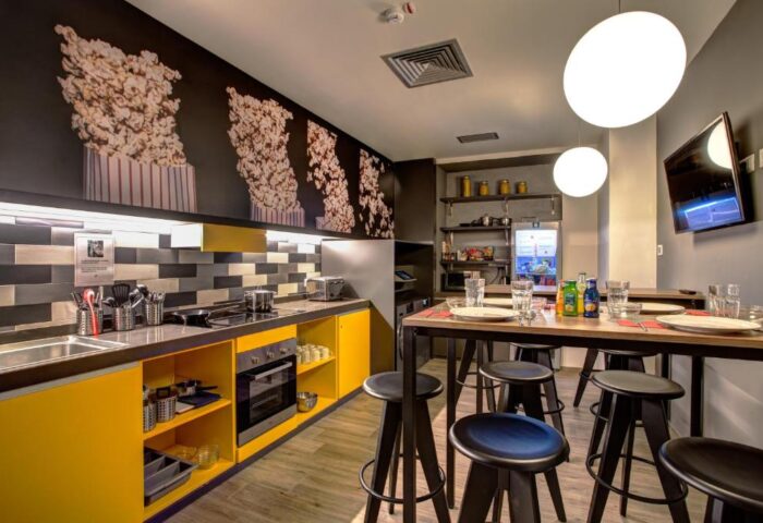 MEININGER Roma Termini gay-friendly hotel kitchen. Yellow cabinets, popcorn wall art, dining table with stools, TV. Modern design.