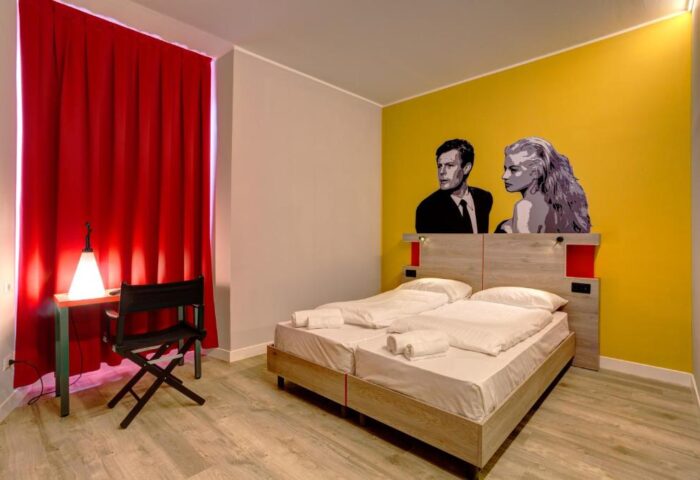 MEININGER Roma Termini gay-friendly hotel room. Double bed with white sheets, desk, chair, red curtains, and a yellow wall with a black and white graphic.