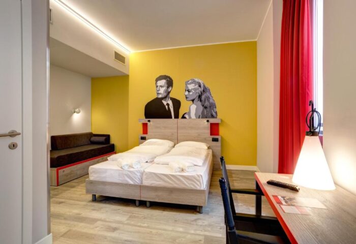 MEININGER Roma Termini gay-friendly hotel room. Double bed, yellow wall with mural, desk, and sofa. A comfortable stay in Rome for gay travelers.