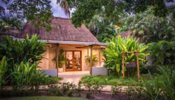 Mahekal Beach Front Resort and Spa gym. Gay-friendly hotel with thatched roof, lush greenery, and exercise equipment visible through glass doors.