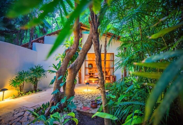 Lush garden view of a bungalow at Mahekal Beach Front Resort and Spa, a gay-friendly hotel. Tropical plants surround a cozy, warmly lit room.