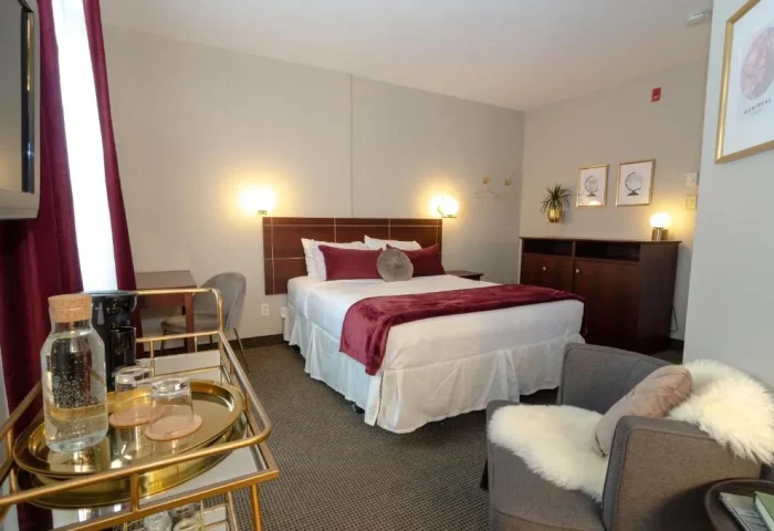 Maison Cartier Montreal gay-friendly hotel room. Bed with burgundy accents, chair with fur throw, and gold bar cart. Upscale, cozy ambiance.
