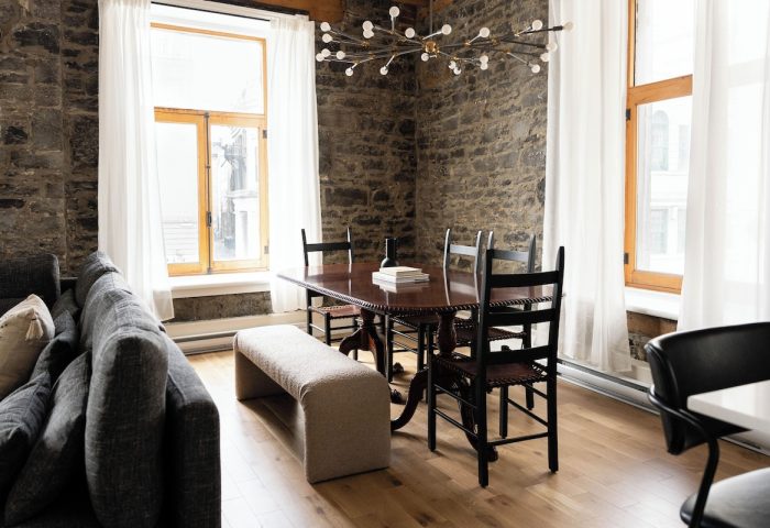 Maison Place Royale interior. Gay-friendly hotel dining area with stone wall, wood table, chairs, bench, and modern chandelier. Luxury In Transit.