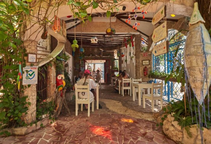 Maison Tulum's gay-friendly hotel restaurant. Open-air dining with white tables/chairs. Lush greenery, colorful decor, and unique artistic touches.