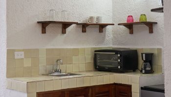 Maison Tulum gay-friendly hotel kitchenette. Sink, microwave, coffee maker, and mini-fridge on the counter. Shelves with glasses and mugs.