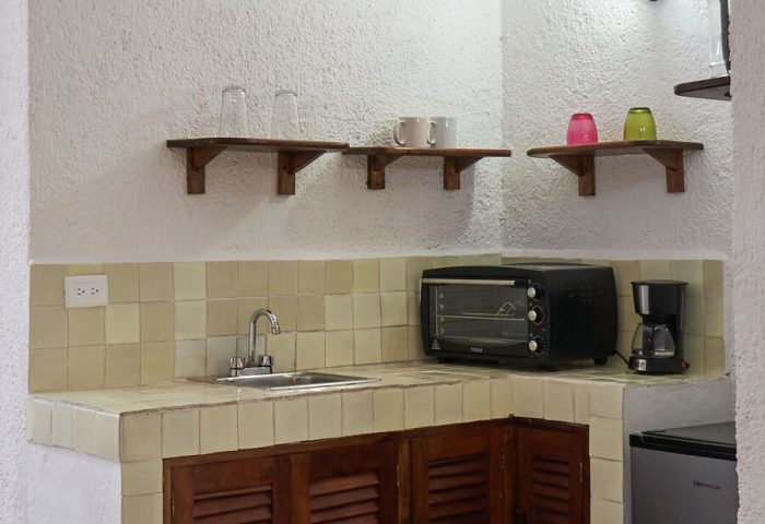 Maison Tulum gay-friendly hotel kitchenette. Sink, microwave, coffee maker, and mini-fridge on the counter. Shelves with glasses and mugs.