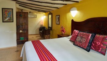 Maison Tulum gay-friendly hotel room. Interior with bed, colorful pillows, yellow wall, and rustic wood accents. Gay travel lodging.