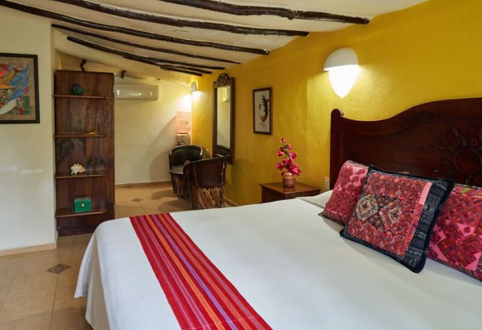 Maison Tulum gay-friendly hotel room. Interior with bed, colorful pillows, yellow wall, and rustic wood accents. Gay travel lodging.