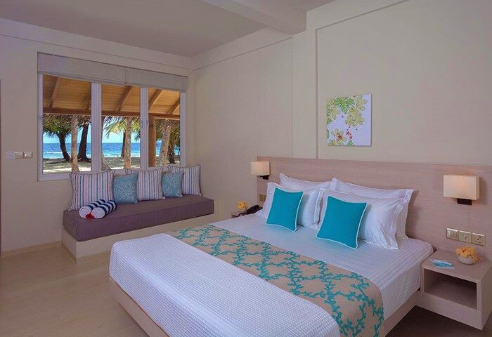 Malahini Kuda Bandos Resort gay-friendly hotel room. Bed with turquoise accents, window seat with ocean view. Relaxing Maldives travel.