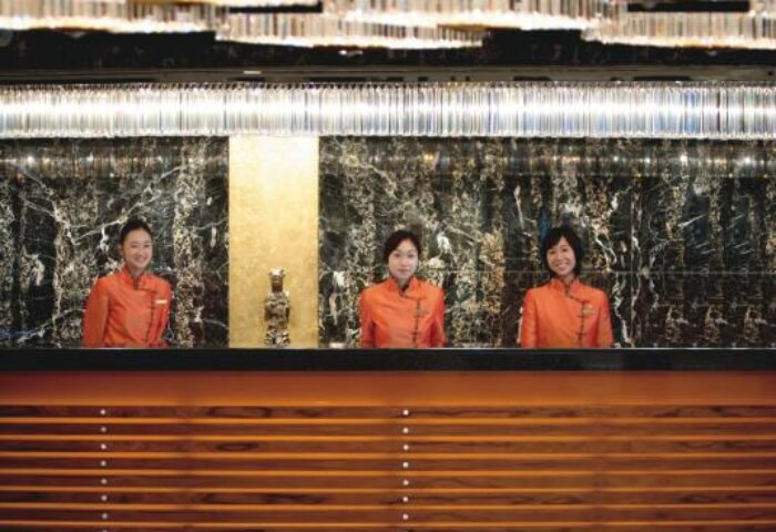 Reception at the gay-friendly Mandarin Oriental Hong Kong hotel. Staff in orange uniforms stand behind a wooden counter under a crystal chandelier.