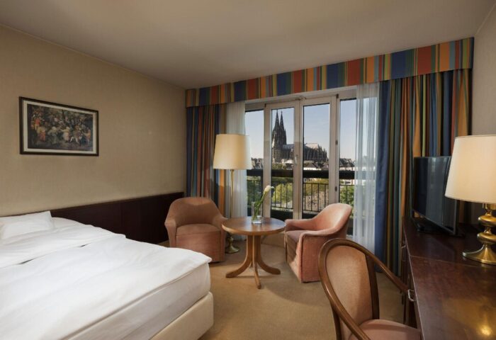 Maritim Hotel Koeln room with Cologne Cathedral view. Gay-friendly hotel room interior with bed, chairs, desk, and balcony. Cologne travel.