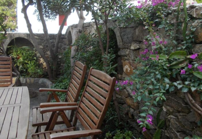 Gay-friendly Matina Hotel seating area. Wooden chairs, stone wall with arches, and bougainvillea. Ideal for gay men's travel and relaxation.