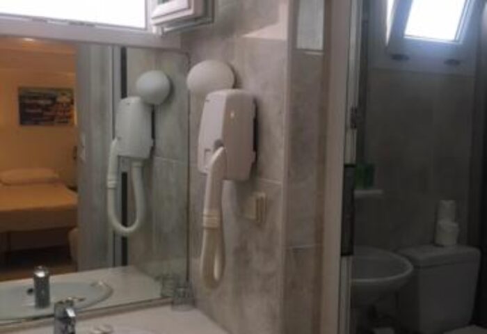 Bathroom interior at Matina Hotel, a gay-friendly hotel. Sink, mirror, hair dryer, toilet, and window visible. Gay travel accommodations.