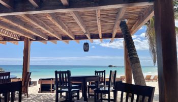 Beachfront dining at Maya Tulum By G Hotels, a gay-friendly hotel. Tables and chairs on the sand under a wooden pergola, ocean view.