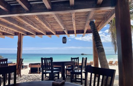 Beachfront dining at Maya Tulum By G Hotels, a gay-friendly hotel. Tables and chairs on the sand under a wooden pergola, ocean view.