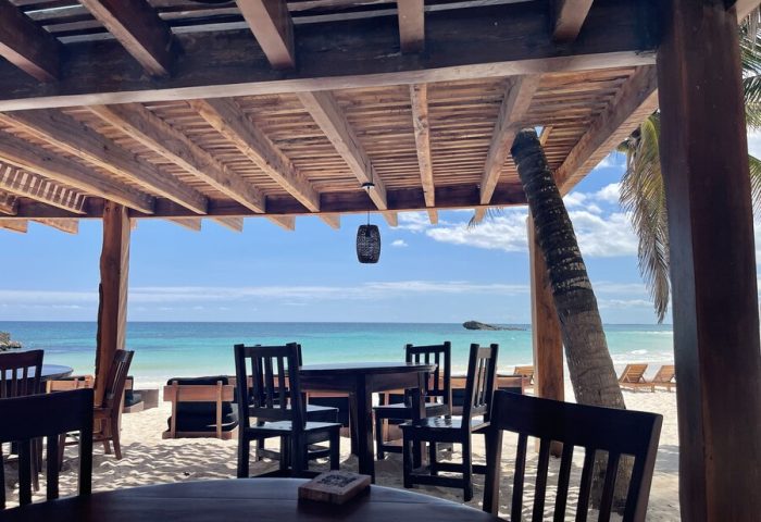 Beachfront dining at Maya Tulum By G Hotels, a gay-friendly hotel. Tables and chairs on the sand under a wooden pergola, ocean view.