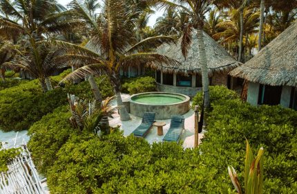 Maya Tulum By G Hotels: Gay-friendly hotel view with thatched-roof cabanas, pool, lounge chairs, and lush greenery. Tropical gay travel.