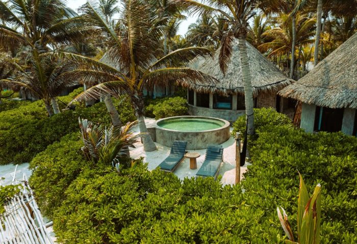 Maya Tulum By G Hotels: Gay-friendly hotel view with thatched-roof cabanas, pool, lounge chairs, and lush greenery. Tropical gay travel.