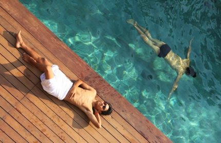 Gay hotel: Man sunbathing by pool, another swimming. Mens resort & spa, gay-friendly travel. Relaxing vacation at a gay hotel.