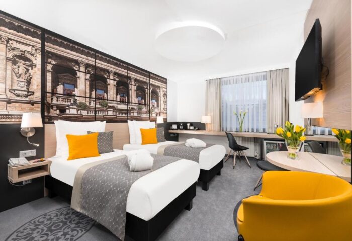 Mercure Budapest City Center Hotel room. Gay-friendly hotel with twin beds, yellow accents, desk, and city view wallpaper. Modern, comfortable stay.