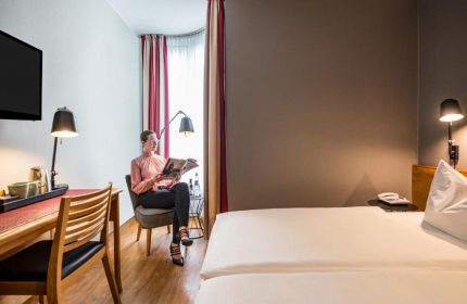 Gay-Friendly Mercure Hotel Berlin Zentrum room. Woman reading. Desk, chair, bed, and lamps. Modern decor. Gay travel lodging.