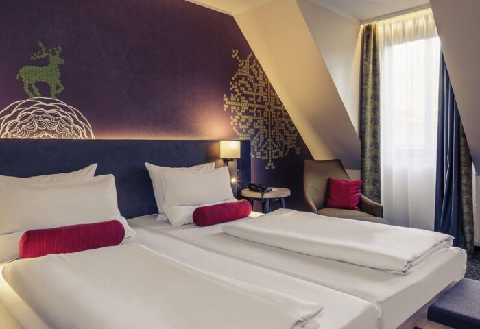 Gay-friendly Mercure Munich City Center Hotel room. Twin beds, purple accent wall with deer design, chair, and window. Comfortable gay travel stay.