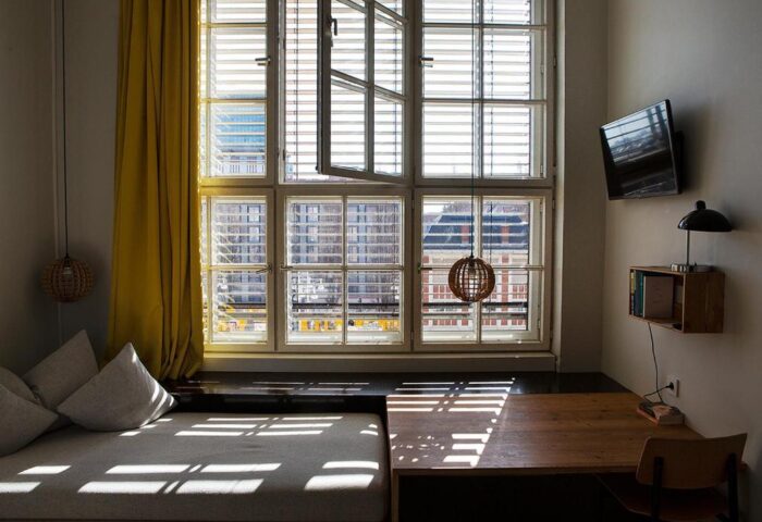 Michelberger Hotel room. Daybed by a window with yellow curtain. Desk, chair, TV, and lamp. Gay-friendly hotel interior.