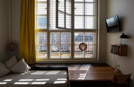 Michelberger Hotel room. Daybed by a window with yellow curtain. Desk, chair, TV, and lamp. Gay-friendly hotel interior.