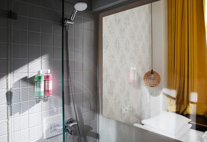 Michelberger Hotel bathroom. Shower with gray tiles and bottles, patterned wallpaper, and a round lamp. Gay-friendly hotel for gay travelers.