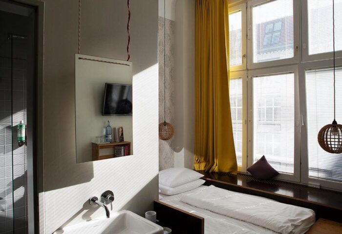 Michelberger Hotel room: single bed by window with yellow curtain. Mirror reflects TV and bathroom sink. Gay-friendly hotel for men's travel.