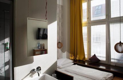 Michelberger Hotel room: single bed by window with yellow curtain. Mirror reflects TV and bathroom sink. Gay-friendly hotel for men's travel.