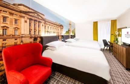 Millennium Gloucester Hotel London room. Gay-friendly hotel with twin beds, red chair, and Buckingham Palace mural. Ideal for gay travel.