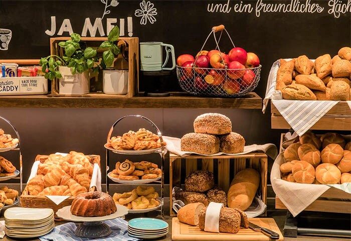 Breakfast buffet at Motel One Berlin-Alexanderplatz, a gay-friendly hotel. Features pastries, bread, fruit, and a chalkboard with German text.