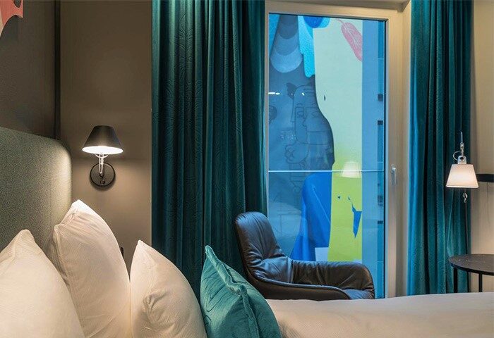 Motel One Berlin-Alexanderplatz room. Gay-friendly hotel with modern design, teal curtains, and a view. Perfect for gay travel in Berlin.