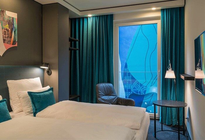 Motel One Berlin-Alexanderplatz room. Gay-friendly hotel with modern design, teal curtains, bed, chair, and city view. Ideal for gay travel.