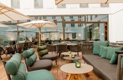Outdoor seating area at Motel One Koln-Neumarkt, a gay-friendly hotel. Tables, chairs, and umbrellas on a wooden deck. Modern design.