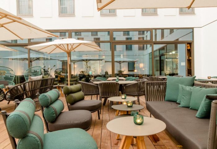 Outdoor seating area at Motel One Koln-Neumarkt, a gay-friendly hotel. Tables, chairs, and umbrellas on a wooden deck. Modern design.