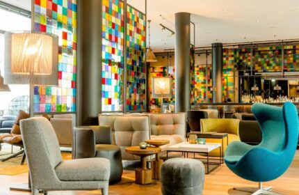Motel One Koln-Neumarkt lobby. Gay-friendly hotel with colorful glass panels, stylish seating, and a modern bar area. Perfect for gay travelers.