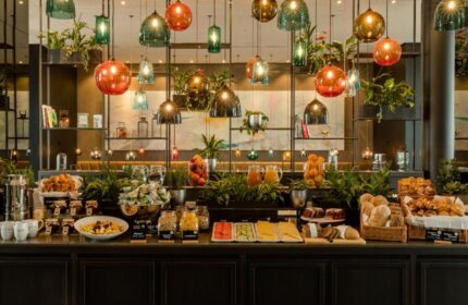 Breakfast buffet at Gay-Friendly Motel One Koln-Neumarkt hotel. Variety of food, drinks, and plants. Unique lighting fixtures.