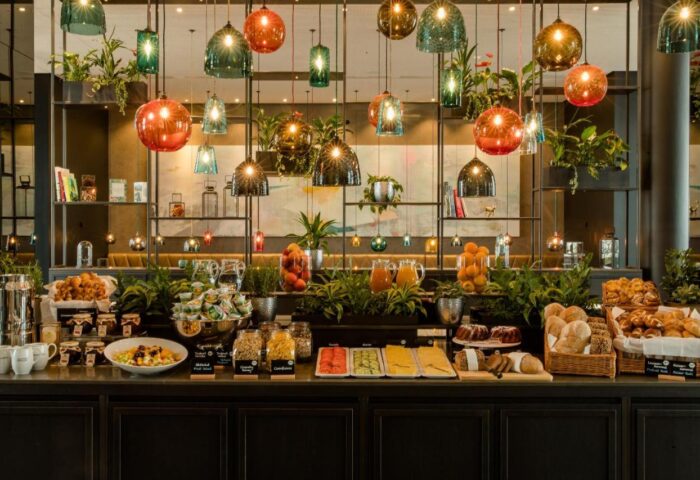 Breakfast buffet at Gay-Friendly Motel One Koln-Neumarkt hotel. Variety of food, drinks, and plants. Unique lighting fixtures.