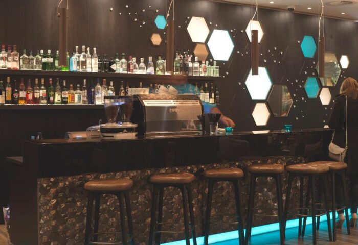Motel One London-Tower Hill bar area. Gay-friendly hotel with stools at the bar, liquor bottles on shelves, and hexagonal lighting.