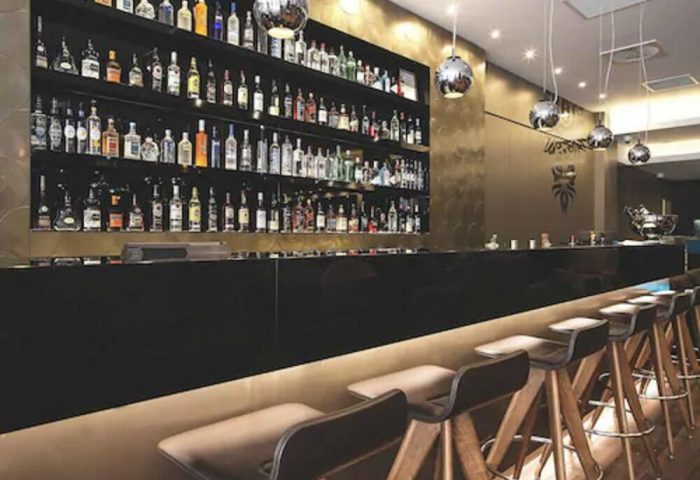 Bar at Motel One Manchester-Piccadilly, a gay-friendly hotel. Shelves stocked with bottles, stools at the bar. Modern design.