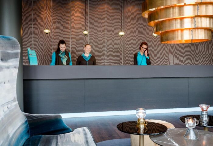 Motel One München Sendlinger Tor reception. Gay-friendly hotel with staff at the front desk. Modern design, warm lighting, and welcoming atmosphere.