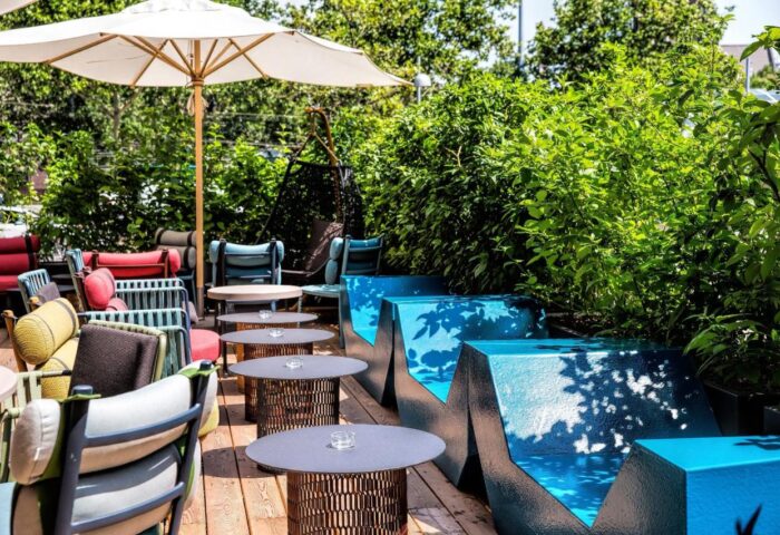 Motel One Wien Westbahnhof's gay-friendly outdoor seating area. Colorful chairs, tables, and blue benches on a wooden deck surrounded by greenery.